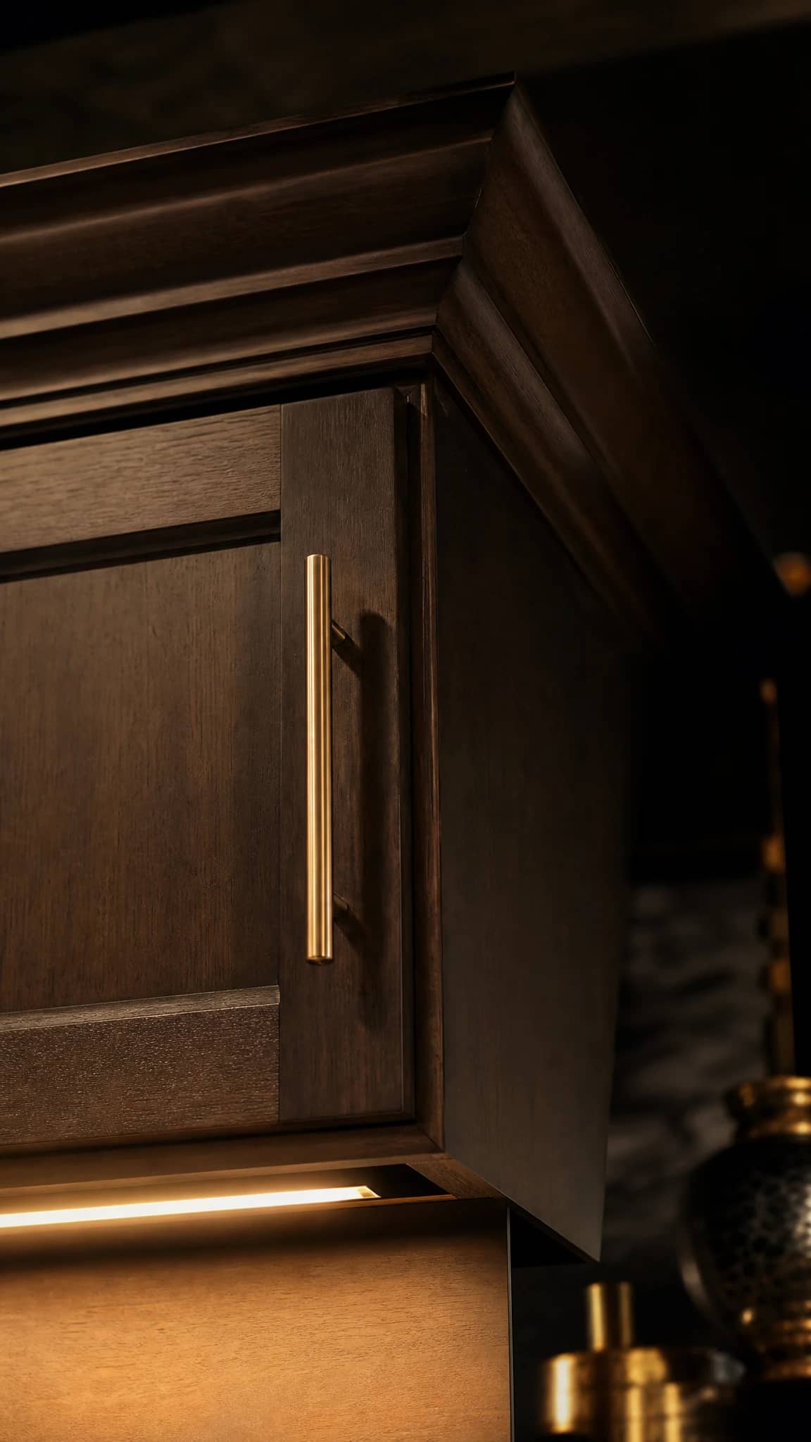 Close view of an espresso shaker door meeting crown moulding, with a brass pull and warm under-cabinet light